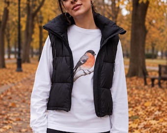 May include: A white long-sleeve shirt featuring a graphic print of a bullfinch bird perched on a branch, layered under a black sleeveless puffer vest with a high collar and black leggings.