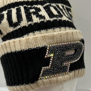May include: Cream and black knit beanie with the word "PURDUE" across the top. The beanie features a black band with a bedazzled "P" logo. A black and white pom-pom sits on top.