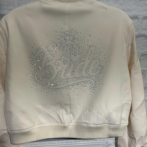 May include: A cream-coloured bomber jacket featuring the word "Bride" in sparkling rhinestones on the back. The jacket has a ribbed collar, cuffs, and hem. A stylish bridal jacket for a wedding.