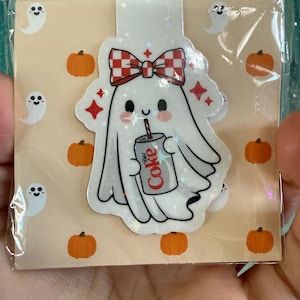 May include: A cute ghost-shaped sticker with a red and white checkered bow, holding a Diet Coke can. The background features small pumpkins and ghost illustrations on a tan background. The sticker is in a clear plastic sleeve.