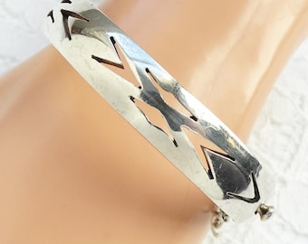 May include: A silver-toned cuff bracelet with a series of cut-out arrow-like shapes. The bracelet is hinged and has a clasp closure. The design is simple and geometric, suitable for everyday wear.