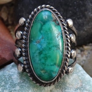 May include: Silver ring with an oval, turquoise-colored stone. The stone is a vibrant mix of green and blue hues, set in a silver band with a twisted rope design around the stone and small silver beads.