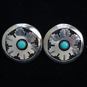 May include: Pair of silver-toned stud earrings. Each earring features a circular design with a turquoise stone center. The outer edge has a geometric pattern. The earrings are displayed against a black background.