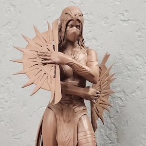 Puede incluir: Escultura marrón de una figura de guerrera femenina, sosteniendo dos objetos en forma de sol. La figura lleva una máscara, armadura y botas. La escultura está sobre una base gris.