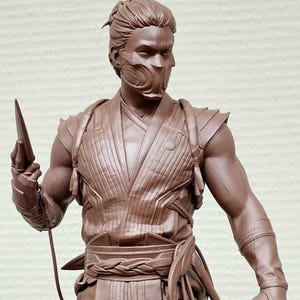 May include: A brown sculpture of a masked warrior figure, holding a bladed weapon. The figure is in a dynamic pose, wearing detailed armor and clothing. The sculpture is mounted on a gray, geometric base.