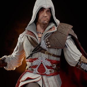May include: A detailed statue of a character from the Assassin's Creed video game series. The figure wears a white and red hooded outfit with brown leather accents, a red sash, and a cape. The statue has a serious expression.