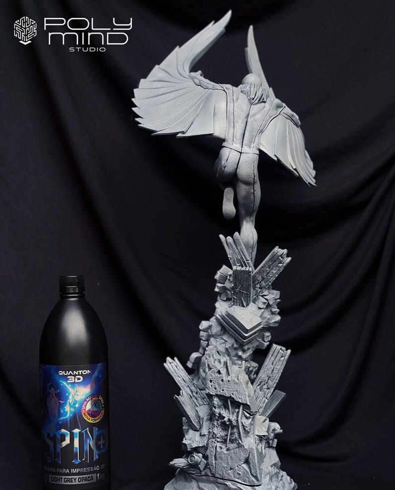 May include: A gray 3D-printed sculpture of a figure with large wings, posed mid-flight, atop a jagged, rocky base. A black bottle with blue and white label is on the left. The sculpture is labeled "POLY MIND STUDIO".
