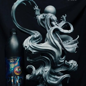 May include: A grey statue of a character with a spherical head, wearing a cape, and holding a book. The statue is on a swirling base. A bottle of "QUANTON 3D" resin is to the left. The Poly Mind Studio logo is in the upper right.