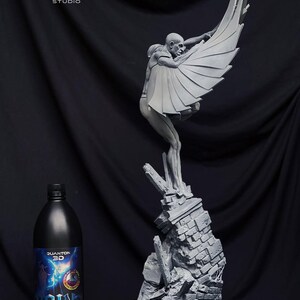 May include: A gray sculpture of a figure with large wings, perched atop a crumbling brick structure. The figure is holding the wings open. A black bottle with text is in the foreground. The sculpture is a detailed art piece.