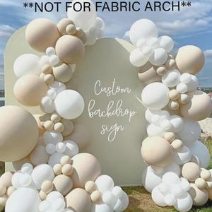 Op de afbeelding: Een aangepast achtergrondbord met een crèmekleurige boog, versierd met een ballonslinger in wit- en beigetinten. Het bord toont "Custom backdrop sign" in sierlijke letters. De tekst "NOT FOR FABRIC ARCH" staat bovenaan.