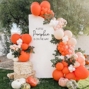 May include: A white sign with the text "A Little Pumpkin Is On The Way" is surrounded by orange, white, and peach balloons. Pumpkins and hay bales are at the base of the sign, creating a festive autumn display.