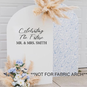 Può includere: Sfondo per matrimoni bianco a forma di arco con la scritta "Celebrating The Future MR. & MRS. SMITH". Il lato destro presenta un motivo floreale blu e bianco. L'arco è decorato con fiori secchi e fiori freschi blu e bianchi.