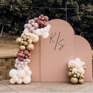 May include: A blush-colored arch backdrop with the initials "K/S" in black script, adorned with a balloon garland. The garland features white, gold, and mauve balloons. A smaller balloon cluster is on the right.