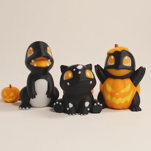 Puede incluir: Tres pequeñas figuras de criaturas estilizadas en negro, naranja y blanco. Una figura es una criatura bípeda negra con vientre blanco, otra es un cuadrúpedo negro con manchas blancas y otra es una criatura negra saliendo de una calabaza.