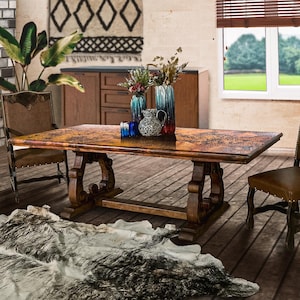 May include: A large, rectangular wooden dining table with ornate, carved legs. The tabletop has a copper-colored finish. Two leather chairs flank the table. Decorative vases and a cowhide rug complete the rustic, elegant dining room setting.