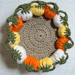 Pode incluir: Uma base de copo de crochê feita à mão com um centro bege e uma borda decorativa de abóboras tridimensionais coloridas. As abóboras são em tons de branco, amarelo e laranja, com caules e folhas verdes. A base de copo foi projetada para proteger as superfícies do calor e da umidade.