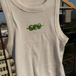 May include: White ribbed tank top with a crew neck and a beaded green dango design. The sleeveless top is hanging on a wooden hanger against a backdrop of a cityscape and a blue sky.