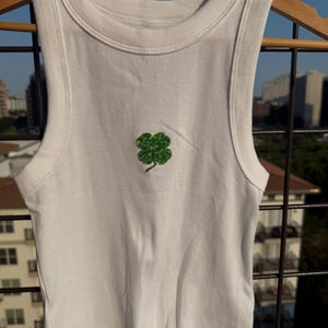 May include: White ribbed tank top with a green glitter four-leaf clover design. The sleeveless top has a round neckline and is hanging on a wooden hanger. The background shows a cityscape and a blue sky.