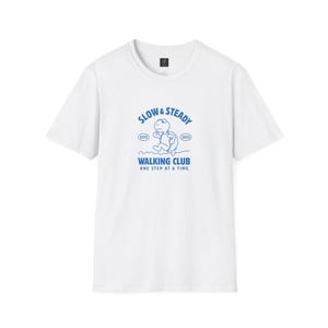 May include: White t-shirt with a blue graphic that reads "SLOW & STEADY WALKING CLUB ONE STEP AT A TIME." The graphic features a turtle walking on a blue line. The shirt has a crew neck and short sleeves.