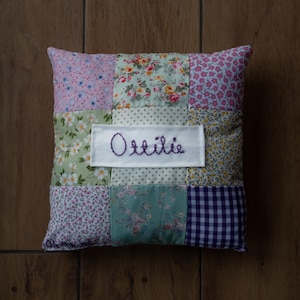 May include: A patchwork cushion featuring floral and patterned fabrics in pink, green, and purple. The name "Ottilie" is embroidered in purple thread on a white rectangular patch in the centre. The cushion is rectangular.