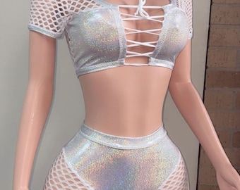 May include: A two-piece silver holographic outfit. The top is a crop top with a lace-up front and white mesh sleeves. The matching shorts have a high waist and white mesh cutouts. The outfit is designed for a party or festival.