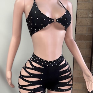 May include: Black two-piece outfit with rhinestone embellishments. The halter top has a ring detail and the shorts feature cut-out designs. The outfit is displayed on a mannequin.