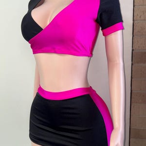 May include: A two-piece outfit featuring a black and hot pink color block design. The top has a wrap-style front with short sleeves. The matching mini skirt has a pink side panel. The outfit is displayed on a mannequin.