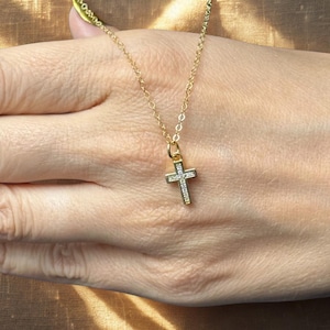 May include: A gold-colored necklace with a small cross pendant. The cross is encrusted with small, clear stones. The necklace is held by a person.