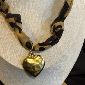 May include: A gold heart pendant necklace. The heart-shaped pendant is attached to a knotted fabric cord with a leopard print pattern in shades of brown and black. The necklace is displayed on a white surface.
