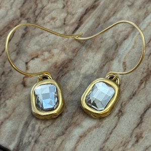 May include: A pair of gold-toned earrings with a rectangular, clear gemstone set in a gold bezel. The earrings have a simple, curved hook design. The background is a mottled, brown and beige surface.