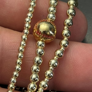 May include: Close-up of a gold baseball-themed necklace. The necklace features a baseball charm with red stitching and is strung with small, round gold beads. The design is a sports-themed jewelry piece.