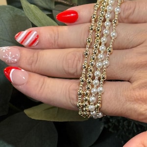 May include: Four beaded bracelets. Two are gold-colored with small round beads, and two are white with pearl-like beads. The bracelets are stacked on a finger. The background is blurred green foliage.