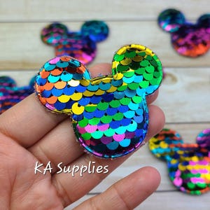 Könnte beinhalten: Regenbogenfarbene, irisierende Pailletten-Mickey-Mouse-Ohren, etwa 5 cm groß. Die Ohren bestehen aus Pailletten, die auf einen Filzgrund genäht sind.