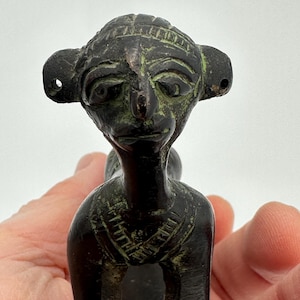 May include: A dark bronze-colored figurine of a stylized human figure with large ears and a detailed face. The figure has a slender neck and appears to be seated. The bronze has a green patina in areas, suggesting age.