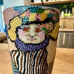 May include: A colorful ceramic vase with a whimsical face design. The vase features a portrait with a floral headpiece, a purple scarf, and a striped pattern. The color palette includes pink, blue, yellow, and brown.