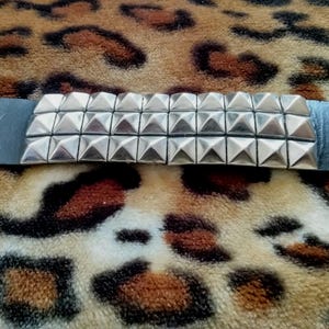 Może przedstawiać: Czarna skórzana bransoletka z dwoma rzędami srebrnych ćwieków w kształcie piramid. Bransoletka ma dwa zapięcia na zatrzask i dwie metalowe przelotki. Bransoletka jest prezentowana na powierzchni z nadrukiem w cętki.