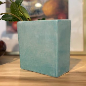 May include: A square, pale blue ceramic vase with a matte finish. The vase holds green leafy branches. A wooden bowl and a framed picture are in the background. The vase is approximately 10 cm tall.
