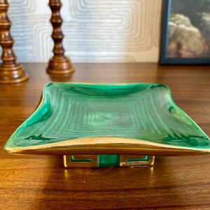 May include: A square, green decorative dish with a gold rim and base. The dish has a malachite-like pattern and is set on a wooden surface. Two wooden candle holders are in the background.