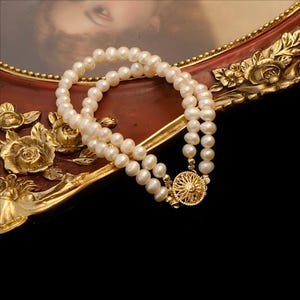 Puede incluir: Collar de perlas de doble hebra con cierre dorado. El cierre tiene un diseño de sol. Las perlas blancas se muestran sobre un marco decorativo rojo y dorado.
