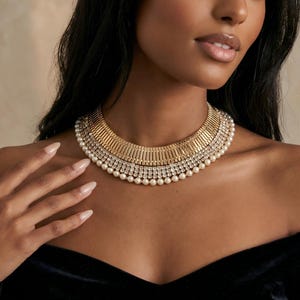 May include: A multi-layered choker necklace featuring a top row of gold-toned vertical bars, a middle row of clear sparkling rhinestones, and a bottom row of small white pearls. The piece sits flush against the neck with a structured design.