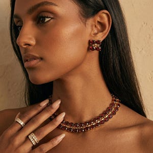 May include: A woman wearing a gold necklace, earrings, and rings. The jewelry features red gemstones and small white stones. The necklace is a double strand. The earrings are square-shaped. The rings are gold with white stones.