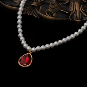 Puede incluir: Un collar de perlas con un colgante de gema roja en forma de lágrima. El colgante está enmarcado en oro y cuelga de un hilo de perlas blancas. El collar se muestra sobre un fondo oscuro.