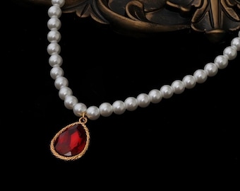 May include: A pearl necklace with a teardrop-shaped, red gemstone pendant. The pendant is framed in gold and hangs from a strand of white pearls. The necklace is displayed against a dark background.