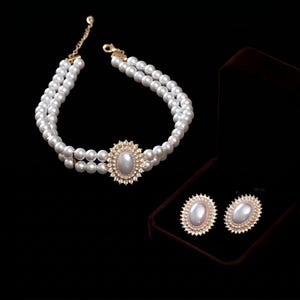 May include: A double-strand pearl choker necklace featuring a central oval cabochon stone surrounded by a halo of small clear crystals, accompanied by a pair of matching oval stud earrings with a similar crystal halo, presented in a dark box.