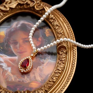 Puede incluir: Un collar de perlas con un colgante en forma de lágrima con una gran gema roja rodeada de perlas más pequeñas y detalles dorados. El collar se exhibe frente a una pintura enmarcada en oro, creando una estética vintage.