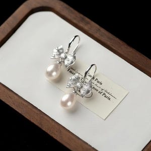 Pode incluir: Brincos de prata elegantes com design de laço, adornados com pequenos cristais transparentes e uma pérola em forma de gota. Os brincos são exibidos em uma superfície branca dentro de uma bandeja de madeira.