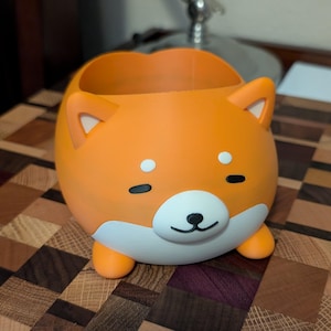 May include: A bright orange Shiba Inu-shaped plant pot with white detailing on the face, ears, and paws. The pot has a rounded body and a large opening for plants. The dog's face has black eyes and a nose.