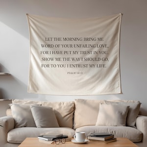 May include: A beige wall tapestry with the text "LET THE MORNING BRING ME WORD OF YOUR UNFAILING LOVE..." is hanging above a beige sofa. A wooden coffee table with books, glasses, and a mug sits in front of the sofa.