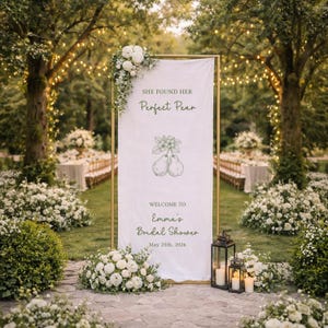 Puede incluir: Un cartel blanco con marco dorado, adornado con arreglos florales, anuncia "She Found Her Perfect Pear" y "Welcome to Emma's Bridal Shower, May 25th, 2026." La escena se desarrolla al aire libre, con vegetaci&oacute;n exuberante y luces de cadena, sugiriendo una boda o evento nupcial.