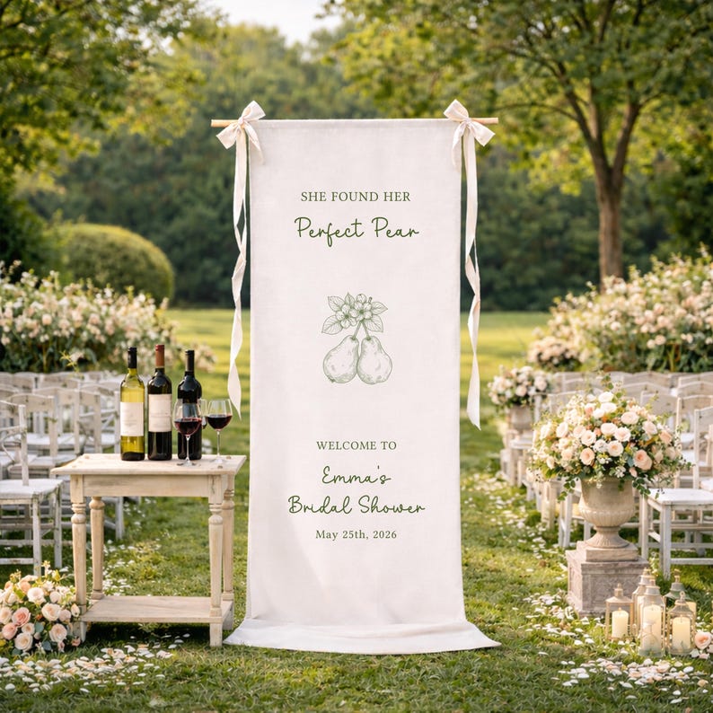Puede incluir: Una pancarta blanca con el texto "She Found Her Perfect Pear" y "Welcome to Emma's Bridal Shower May 25th, 2026". La pancarta est&aacute; decorada con una ilustraci&oacute;n de pera y atada con cintas blancas. Botellas y copas de vino sobre una mesa peque&ntilde;a.